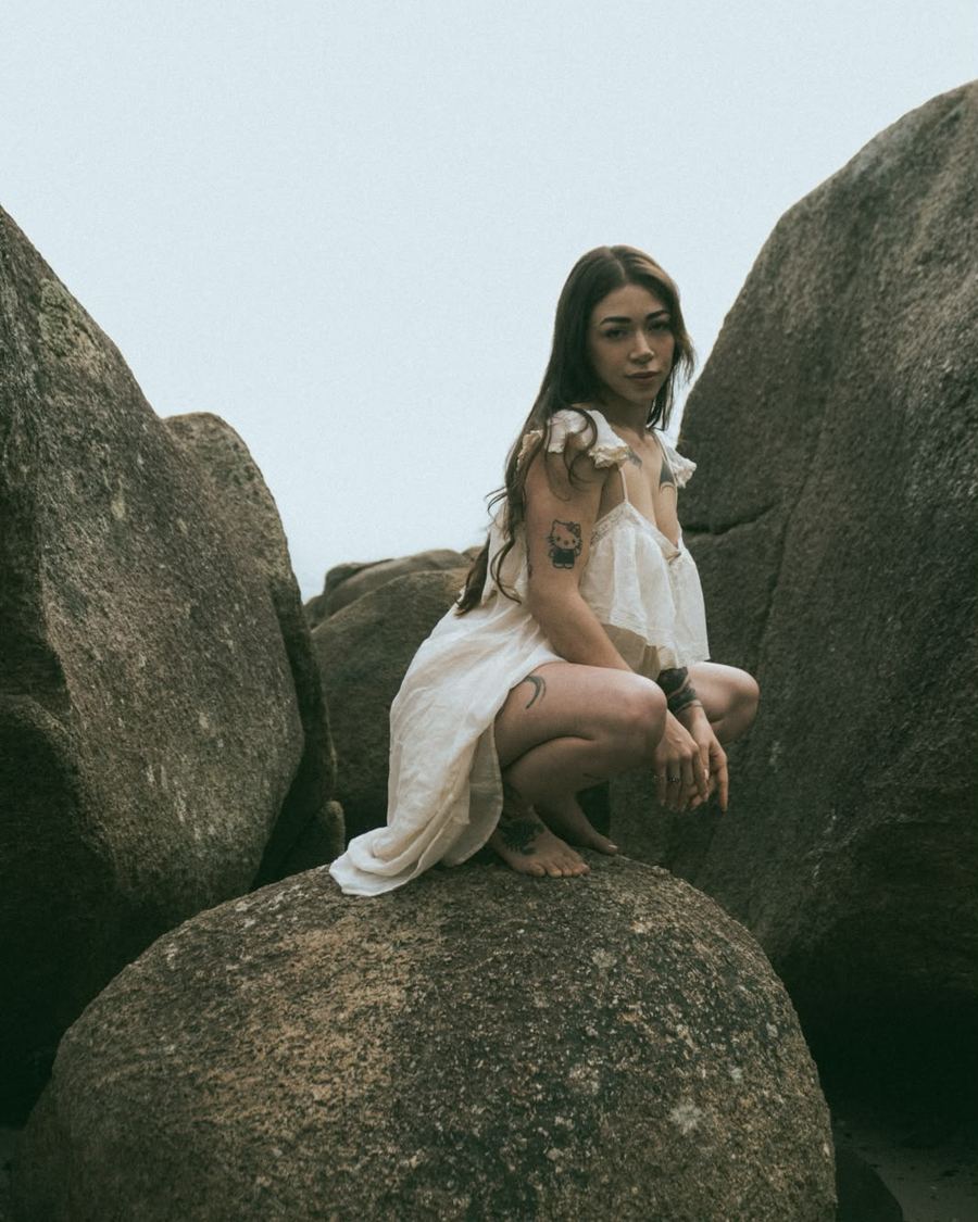 Ana Carla sentada nas pedras da praia, de vestido branco
